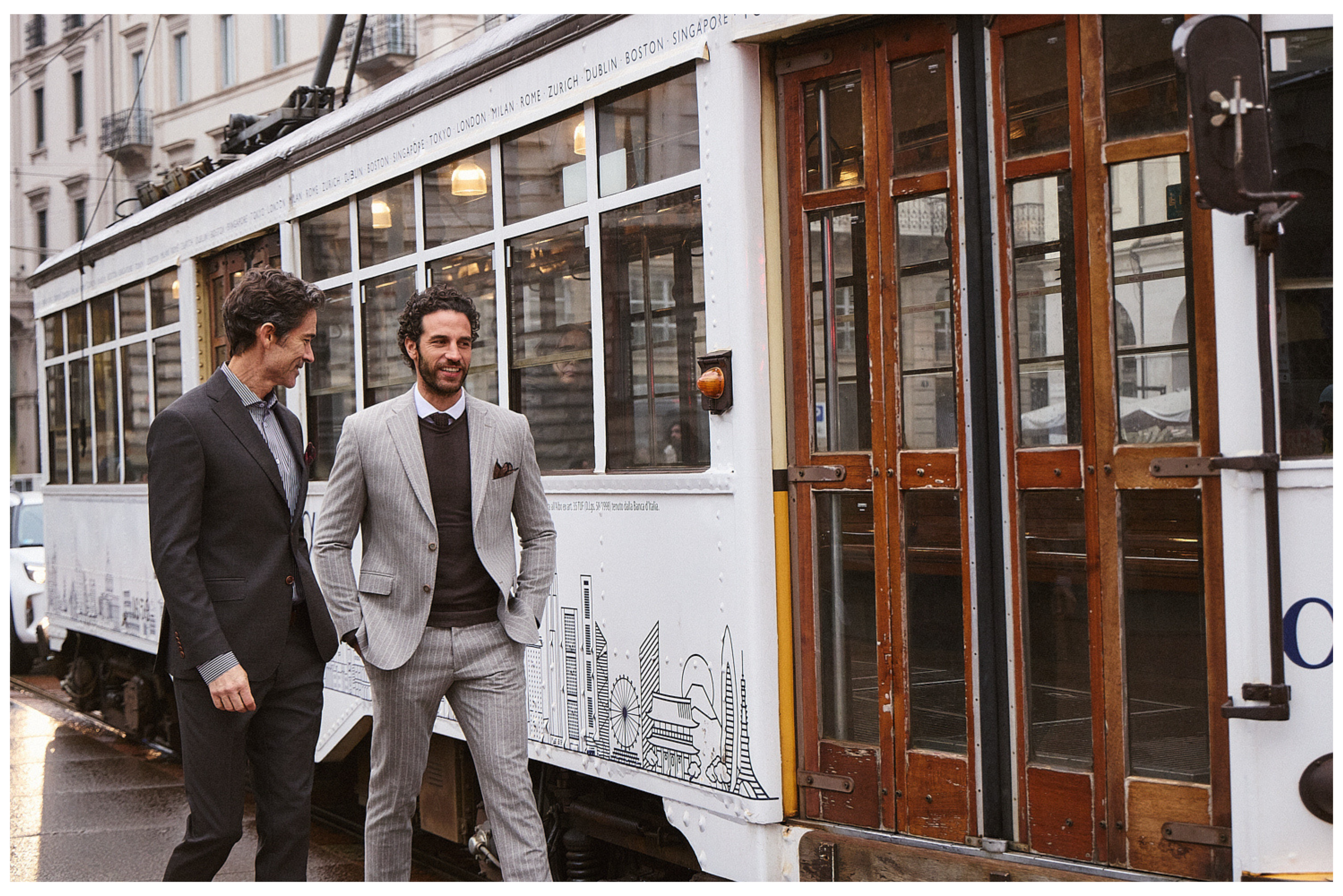 Δύο άνδρες με κοστούμια Portobello's περπατούν δίπλα σε vintage τραμ με ξύλινες πόρτες στους δρόμους του Μιλάνου.