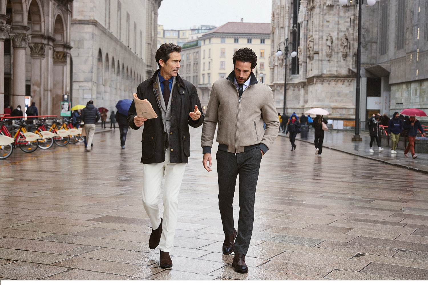 Δύο άνδρες περπατούν και συζητούν σε πλατεία Duomo του Μιλάνου, ντυμένοι με κομψά ρούχα Portobello's. 