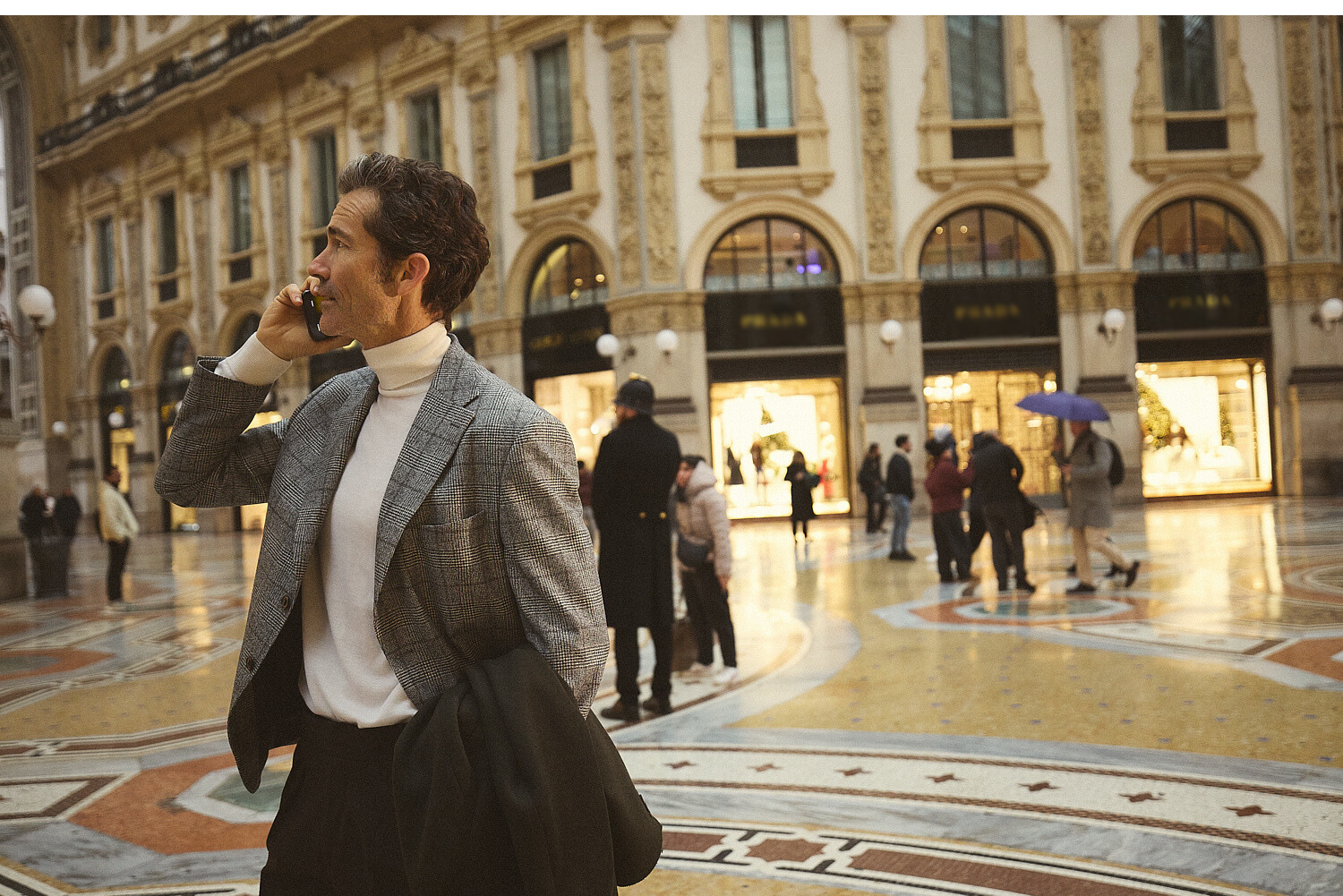 Άνδρας με γκρι σακάκι και λευκό ζιβάγκο Portobello's μιλά στο τηλέφωνο κρατώντας παλτό, στην επιβλητική εμπορική στοά Galleria Vittorio Emanuele II του Μιλάνου.