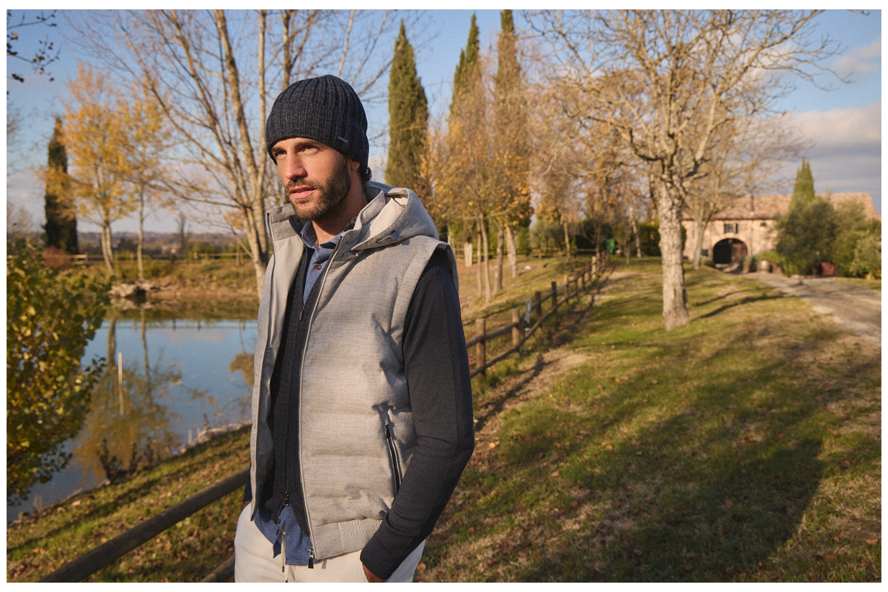 Άνδρας με σκουφί και γκρι γιλέκο Portobello’s στέκεται δίπλα σε λίμνη στο Roncolo, μέσα σε φθινοπωρινό φυσικό τοπίο.
