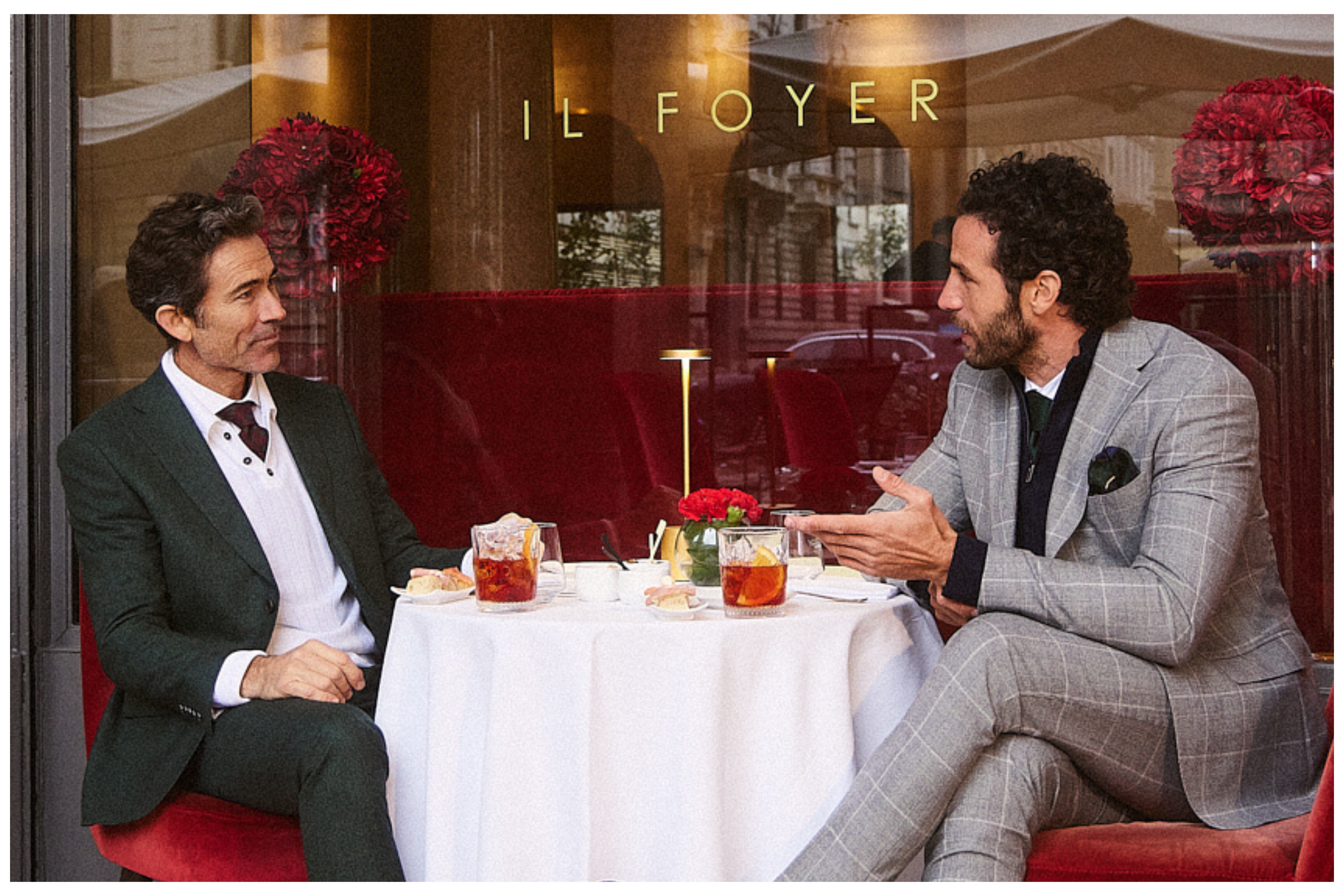 Δύο άνδρες με κοστούμια Portobello's συζητούν σε μικρό τραπέζι με λευκό τραπεζομάντηλο, με ποτά και snacks, έξω από καφέ στο Μιλάνο με την επιγραφή «IL FOYER».
