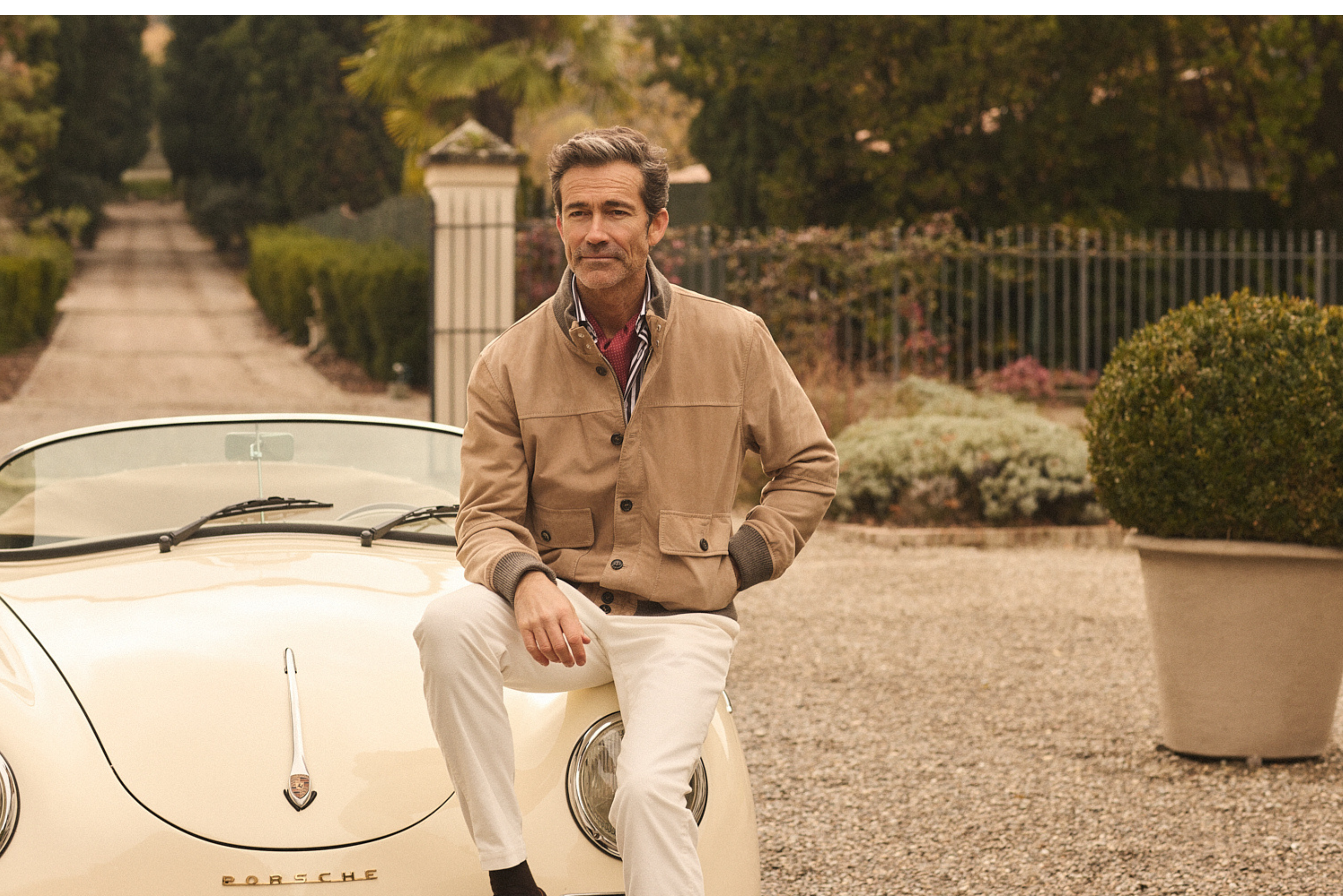 Άνδρας με σύνολο Portobello’s κάθεται σε vintage λευκή Porsche, σε χωμάτινο δρόμο περιτριγυρισμένο από δέντρα, στο πλαίσιο της καμπάνιας Fall–Winter 2026.